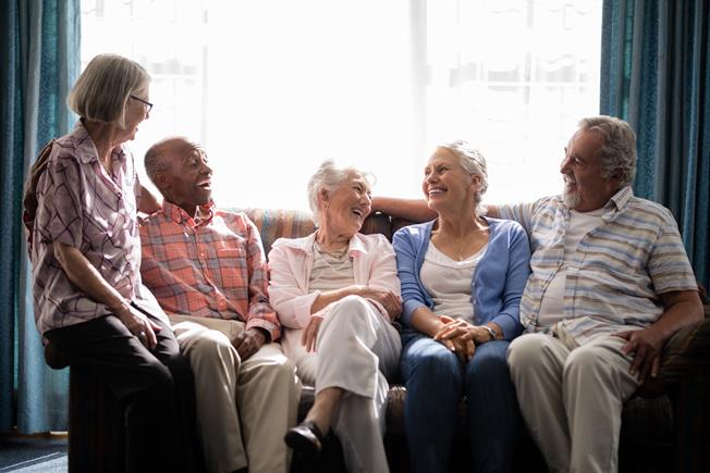A diverse group of five senior friends sitting together on a couch, laughing and talking in a brightly lit room.